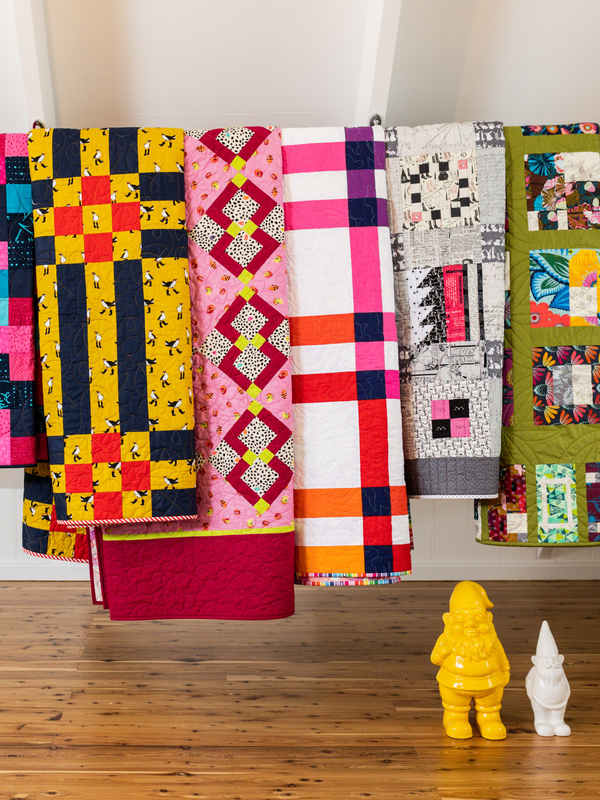 Colourful modern patchwork quilts hanging in a sewing studio, with yellow and white decorative gnomes on the wooden floor.